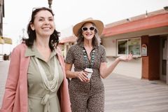Two stylish senior women walking outside, smiling and enjoying coffee on a casual day out.