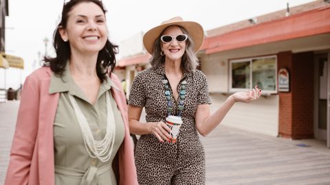 Two stylish senior women walking outside, smiling and enjoying coffee on a casual day out.