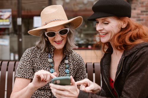 Free Two fashionable women laughing outdoors while using a smartphone, showcasing friendship and style. Stock Photo