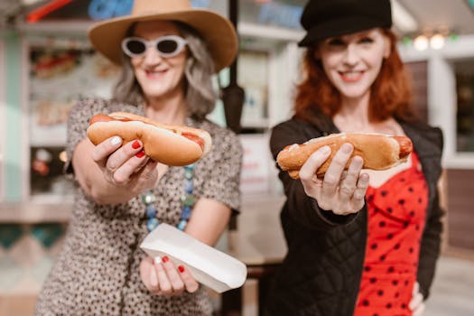 Two women happily holding hotdogs in a vibrant outdoor setting, enjoying casual dining.