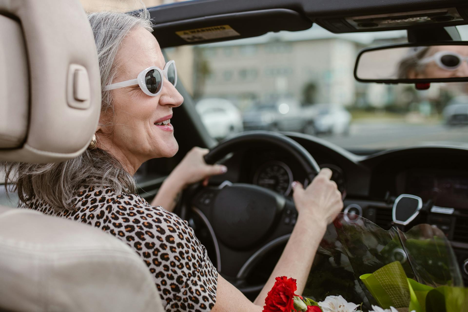 Senior woman driving a convertible, smiling and wearing sunglasses on a sunny day.