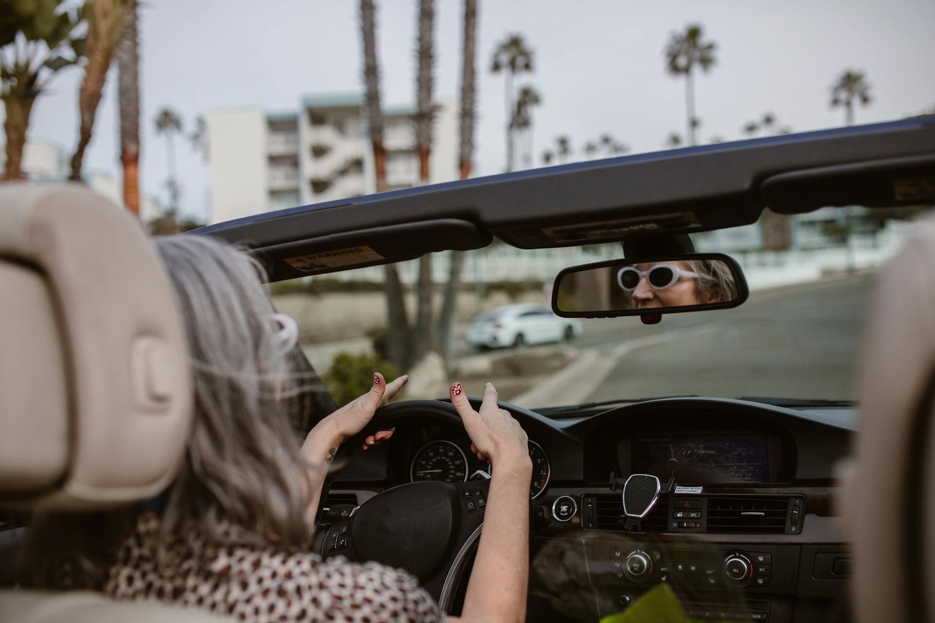 Senior woman enjoying a drive in a convertible with palm trees lining the road.