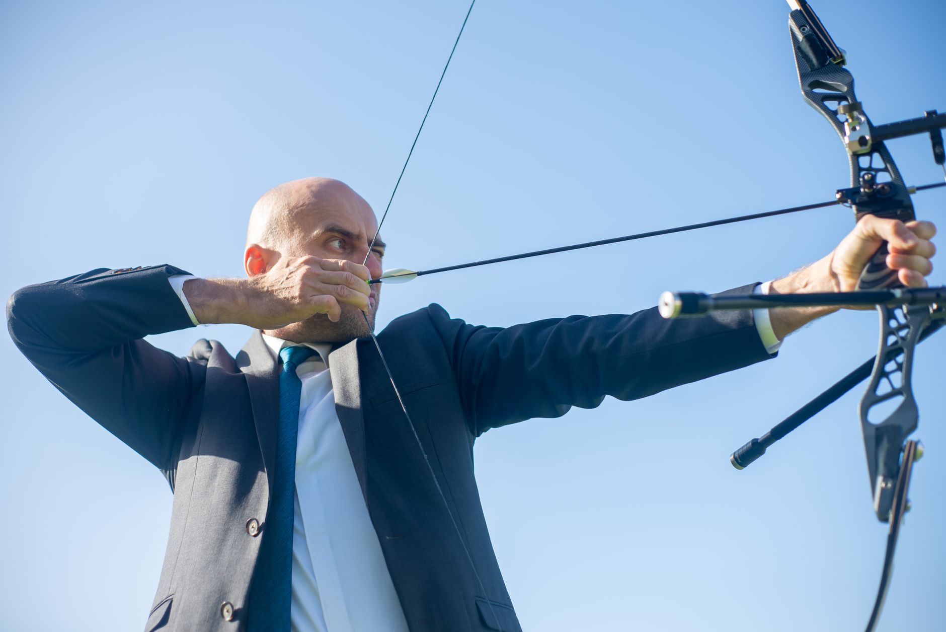 Businessman in a suit aiming a bow and arrow outdoors, exemplifying focus and determination.