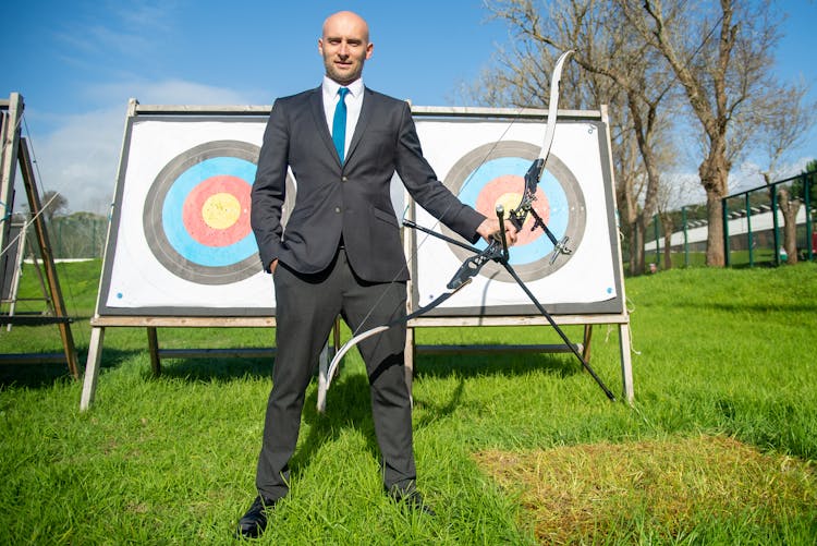 Man In Formal Wear Holding Compound Bow And Arrow Infront Of Archery Target Board