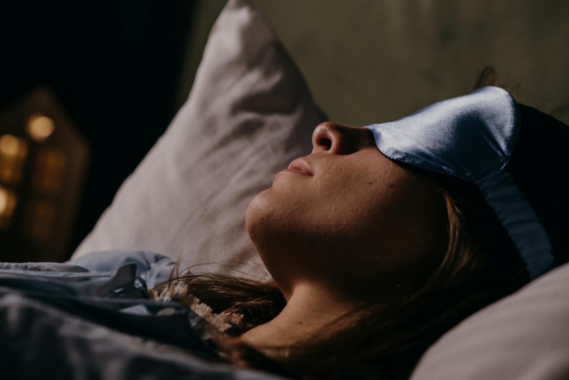 A serene scene of a woman wearing a blue eye mask, sleeping soundly in a cozy indoors setting.