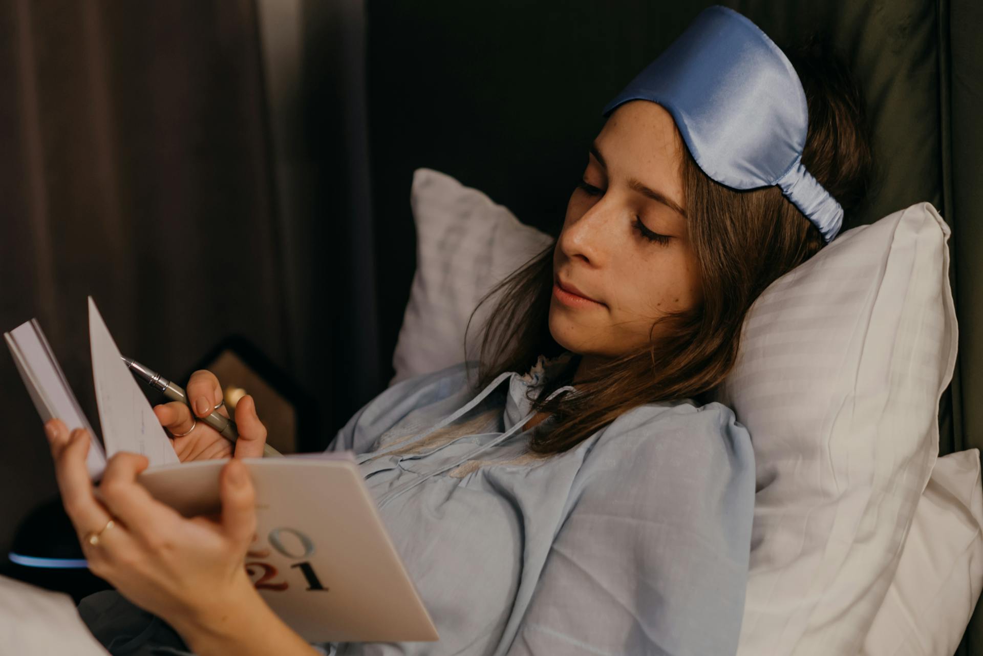 A young woman relaxing and writing in her journal while resting in bed with a sleep mask.