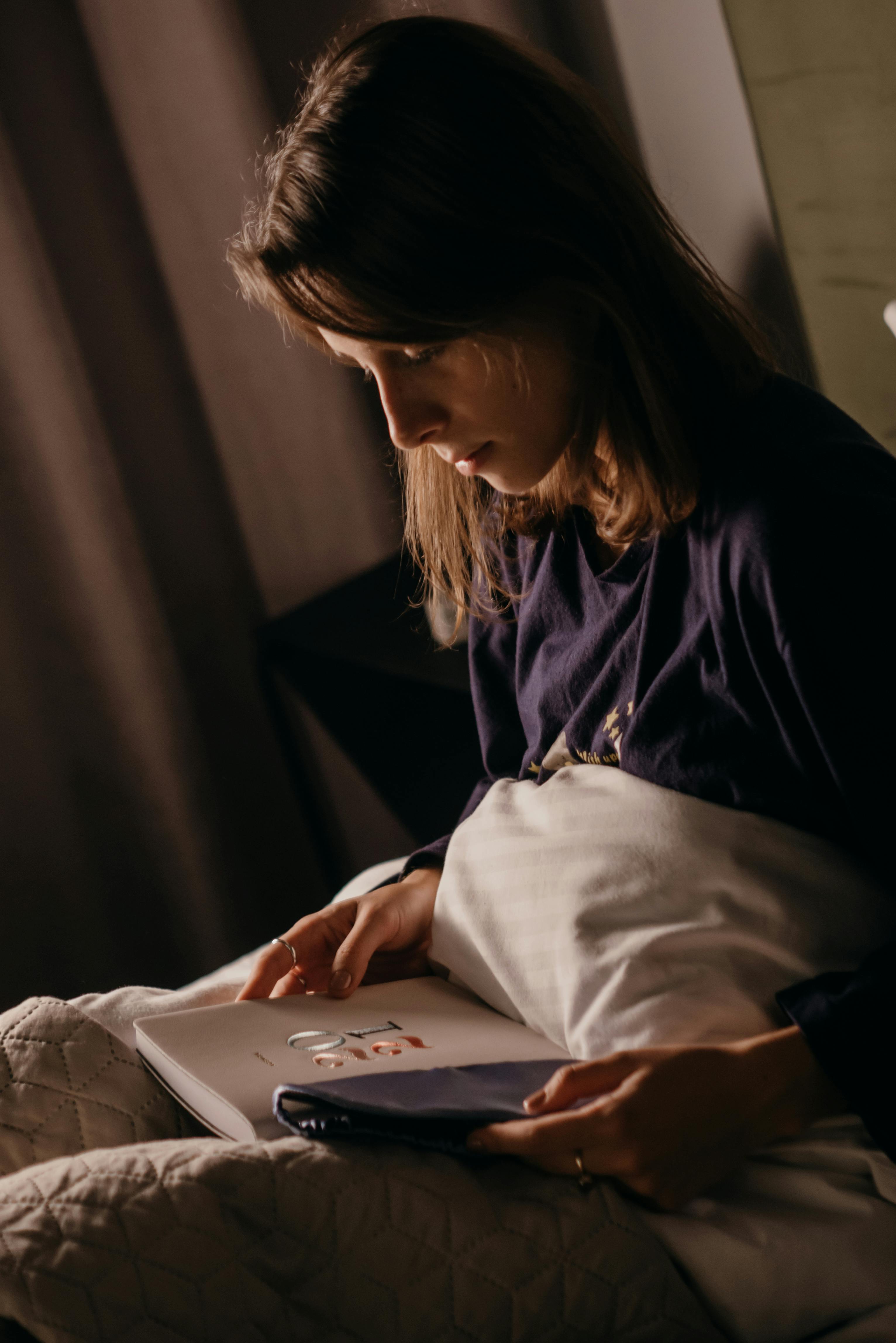 A young woman reading a magazine in a dimly lit cozy room, wrapped in a blanket.