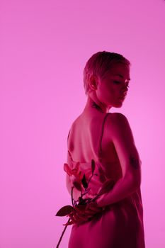 A tattooed woman in a silk dress holds lilies against a pink background, exuding elegance.