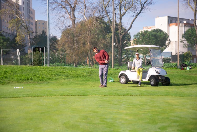 A Woman Sitting On Golf Cart While Looking At The Man Playing Golf