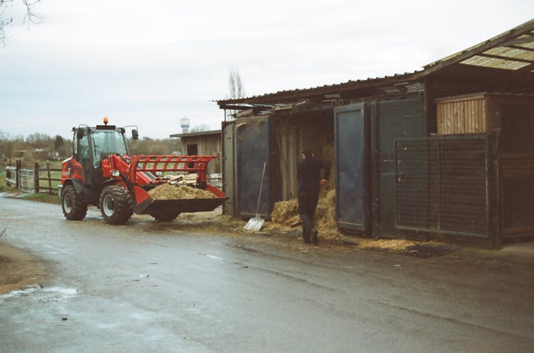 Tractor Parked On Rural Road Near Anonymous Farmer Working Near Barn With Hay