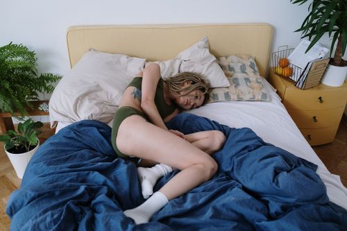 Free Woman lying in bed experiencing abdominal discomfort surrounded by cozy interior. Stock Photo