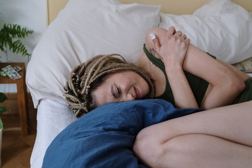Free A woman with dreadlocks lying in bed holding her stomach in pain, possibly due to cramps. Stock Photo