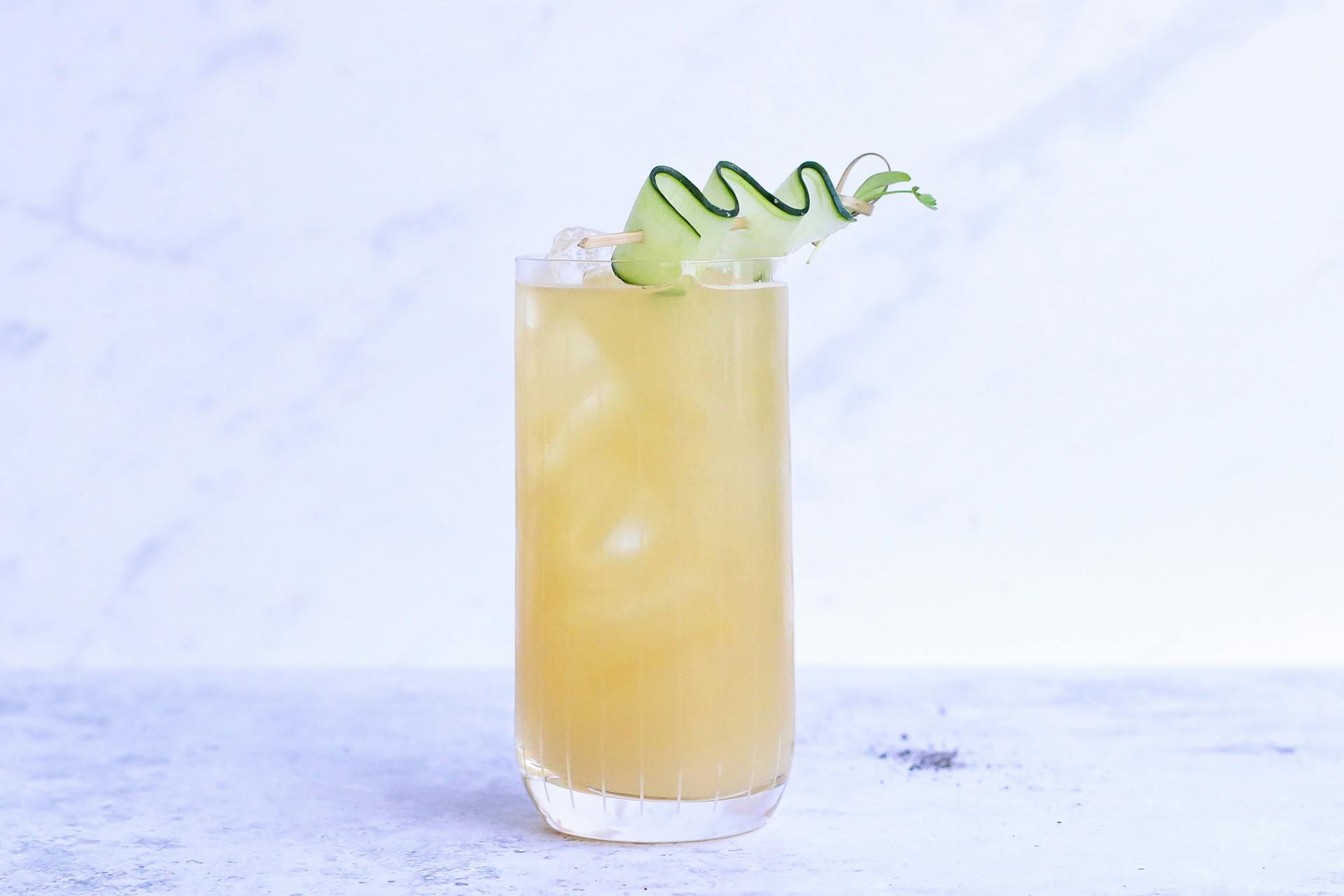 Glass of yummy sour ginger lemonade garnished with cucumber slice and served on light marble table in studio