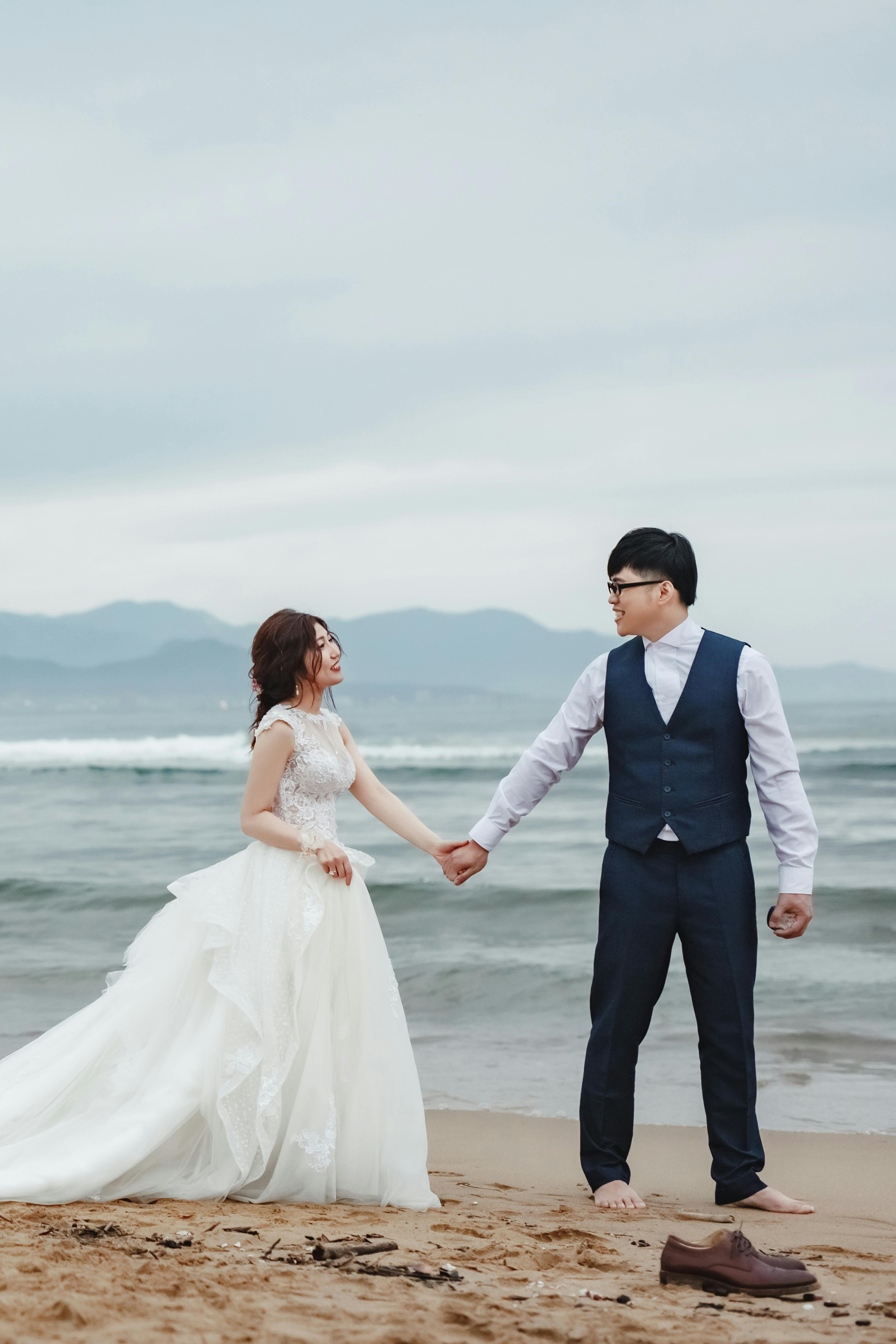 A romantic scene of a couple in wedding attire holding hands on a beach in New Taipei City.