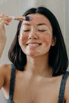Asian woman applying a clay face mask with a brush, showcasing self-care and skincare routine.