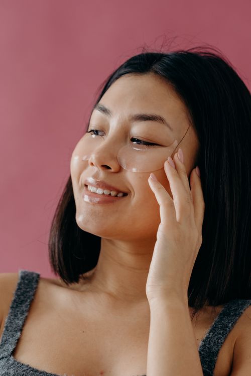 Free Smiling Asian woman applying under eye patches for skin care against a pink background. Stock Photo