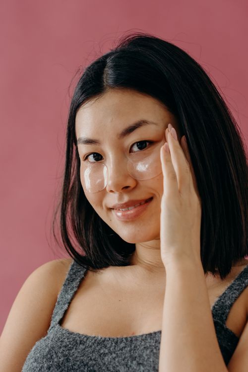 Free Smiling Asian woman applying under eye patches for skin care against a pink backdrop. Stock Photo