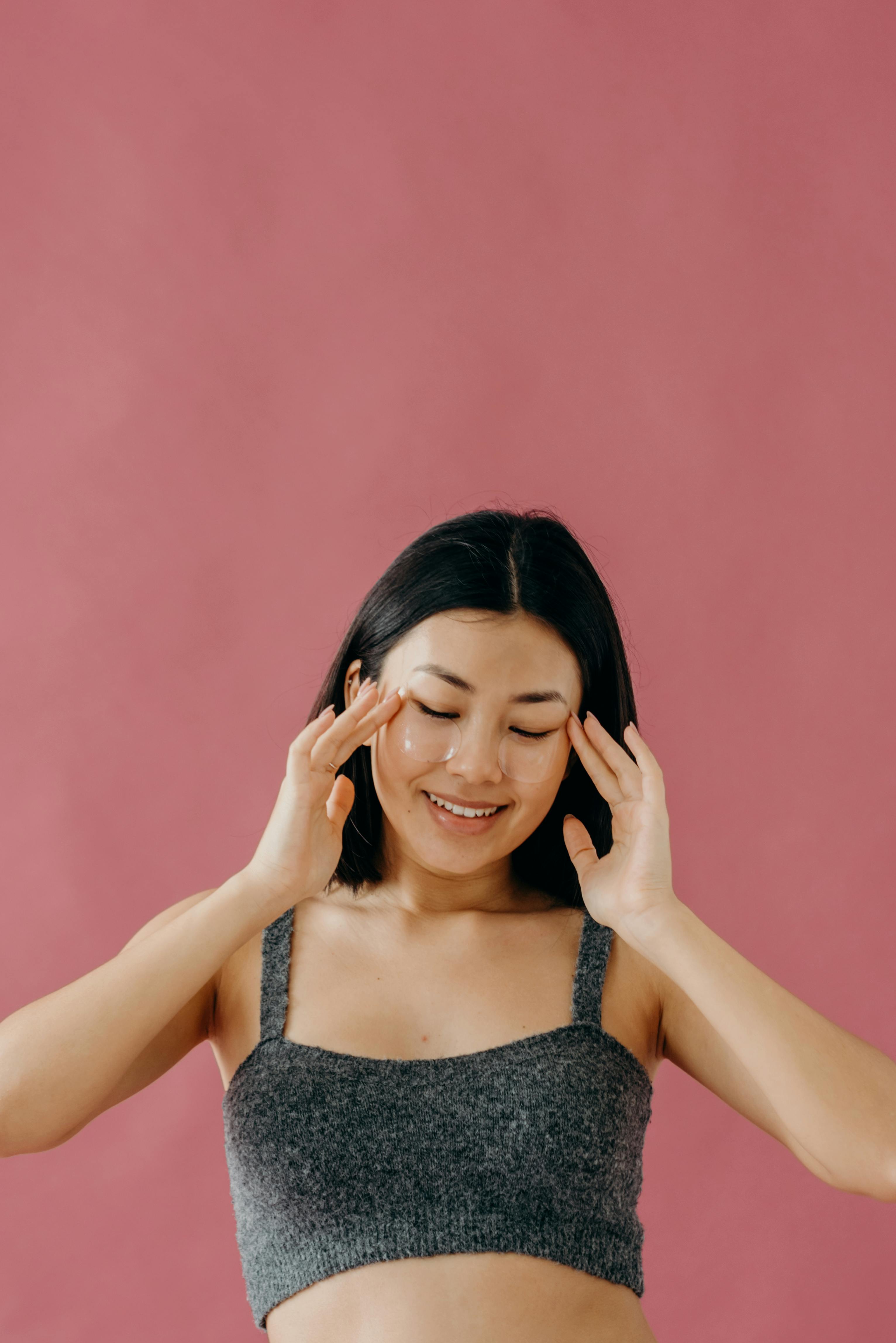 Asian woman enjoying skincare routine with under eye patches on a pink background.