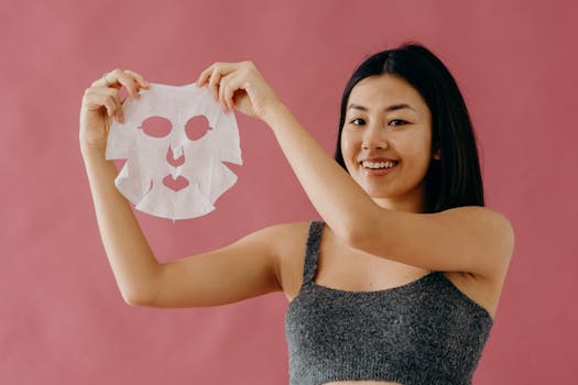 Smiling Asian woman showcasing skincare routine with a sheet mask against a pink backdrop.