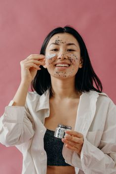 Asian woman smiling while applying an exfoliating face mask for skincare routine.