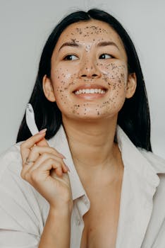 Portrait of a young woman applying a sheet mask with glitters, smiling happily in a studio setting.