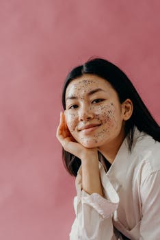 Portrait of a young woman with a glitter facial mask against a pink background, smiling and relaxed.