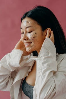Young woman smiling with a glittery facial mask against a pink background, showcasing beauty and skincare.