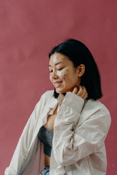 A smiling Asian woman applies a glitter face mask for skincare against a pink background.