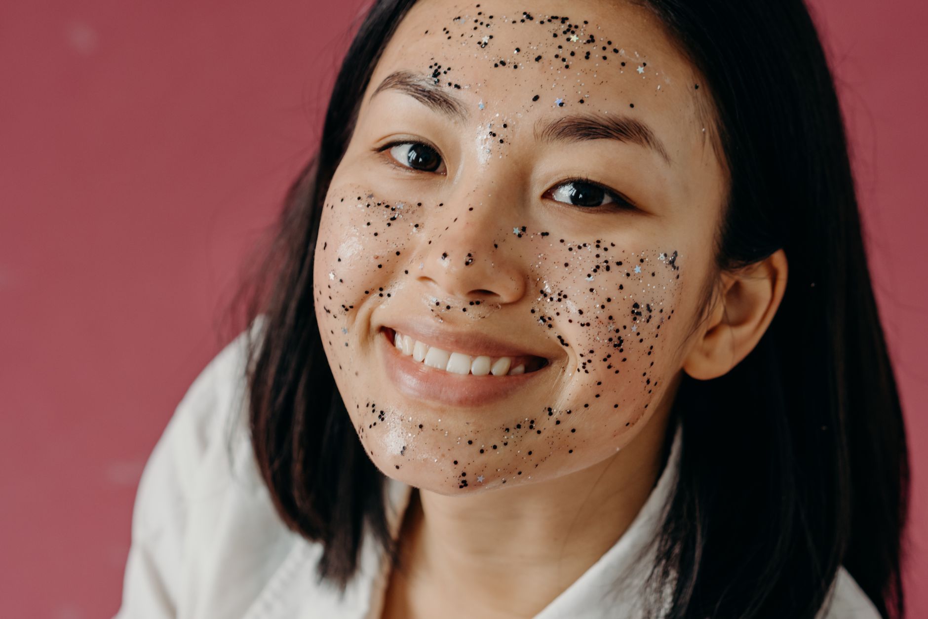 Asian woman smiling with glitter facial mask against pink background, promoting skincare and beauty.