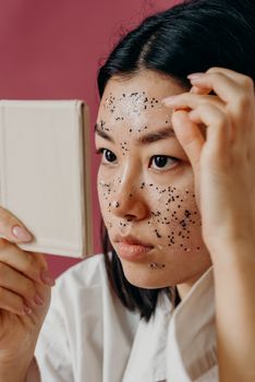 Asian woman examining her facial mask application in a mirror, focusing on skincare.
