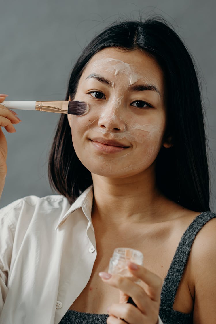 A Woman In Gray Tank Top Applying A Cream On Her Face Using A Brush