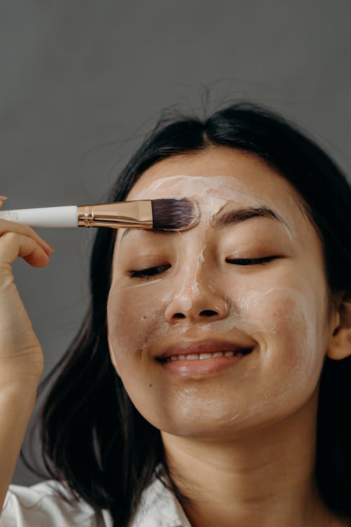 A Smiling Woman Applying a Cream on Her Face Using a Brush