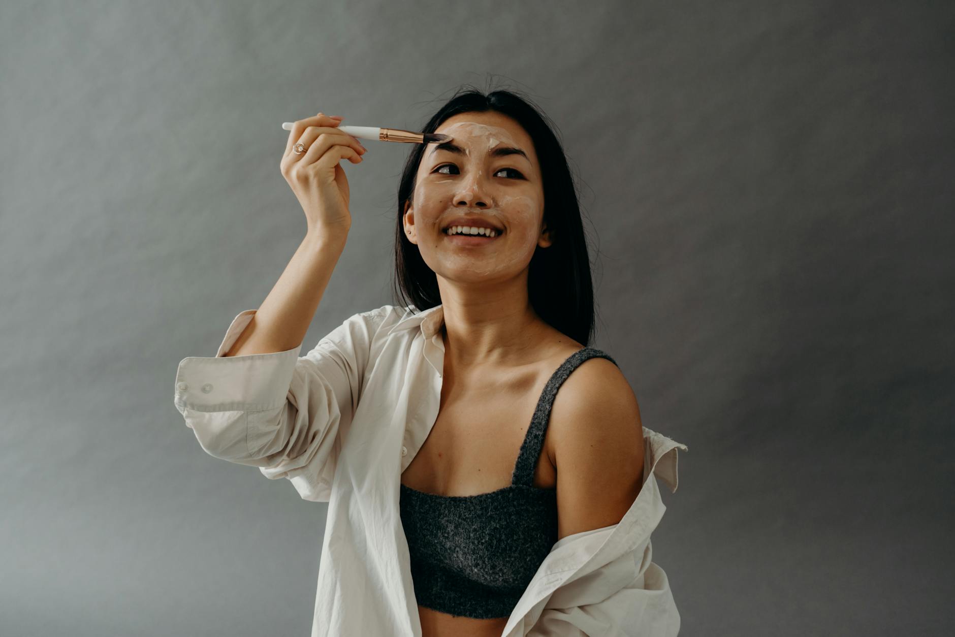 A Woman in White Long Sleeves and Gray Tank Top Smiling while Applying a Cream on Her Face