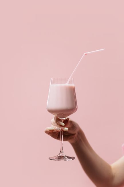 A pastel pink strawberry milkshake in a wine glass held by a hand on a pink background.