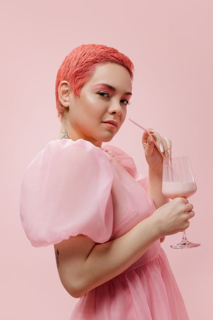 Woman with short pink hair in a pink dress enjoying a strawberry milkshake against a pink background.
