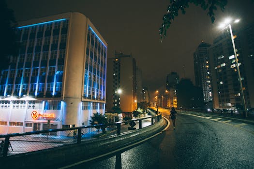A solitary person walks on a wet city street at night, surrounded by illuminated buildings.