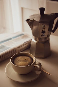 A warm cup of coffee next to a moka pot and a newspaper in a cozy setting.