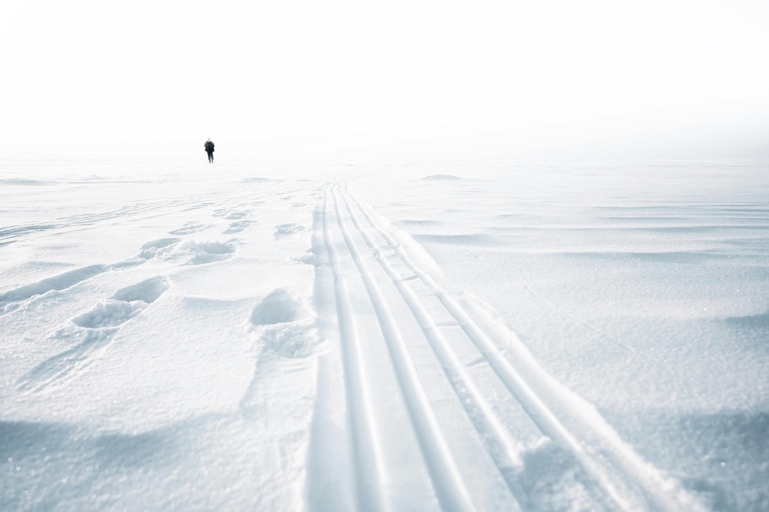 A lone person walks across a vast snow-covered field with tracks in the foreground.