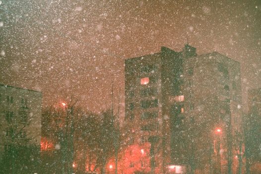A snowy cityscape at night featuring illuminated buildings and snowflakes under streetlights.