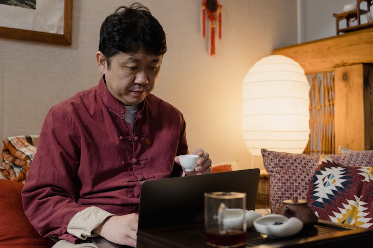 A Man Holding A Teacup While Using His Laptop