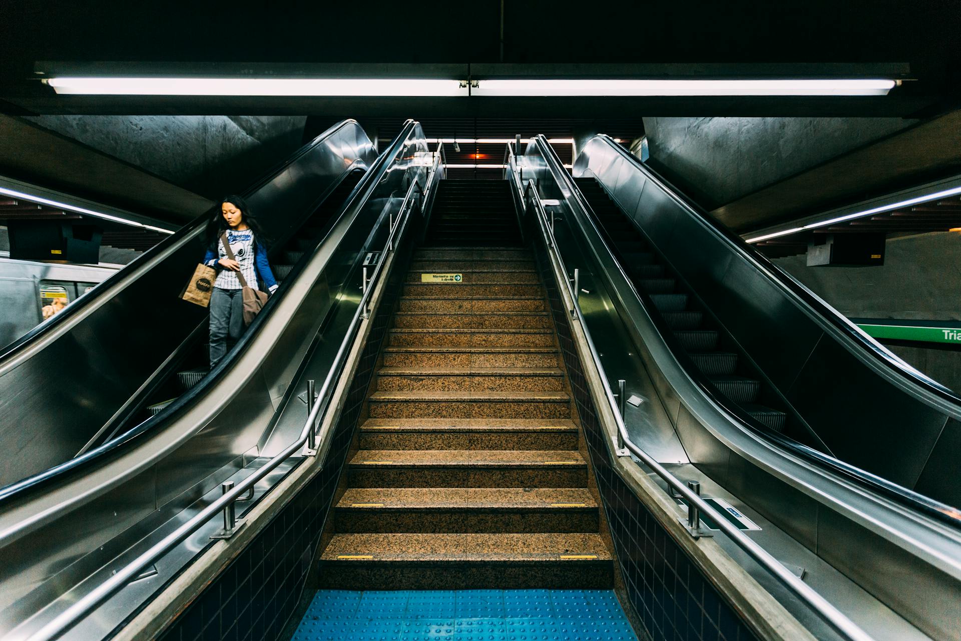 A woman rides escalator next to stairs in a modern subway station setting.