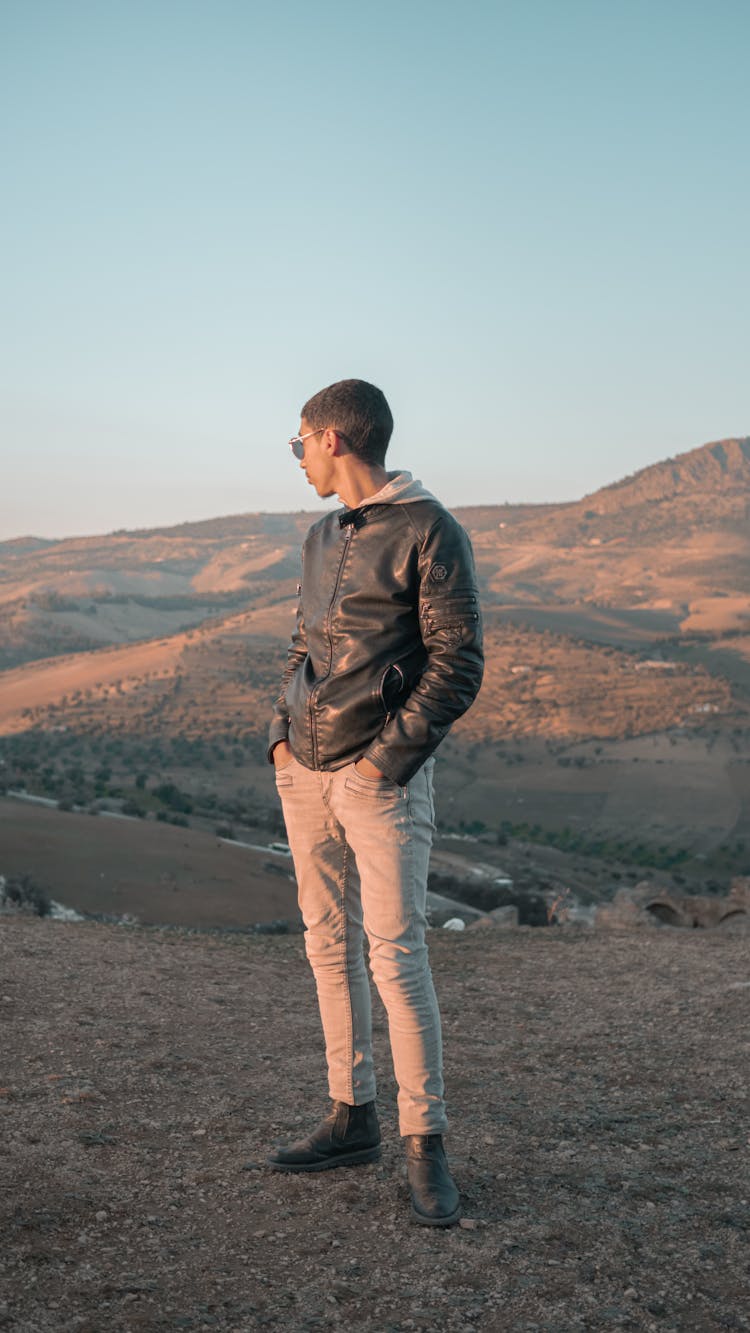 A Man In Black Leather Jacket And Beige Pants Standing On Mountain