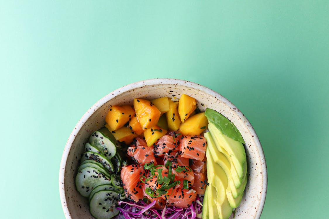 Top view of poke bowl consisting of chopped cucumbers avocado and salmon with mango and blue cabbage placed on green background