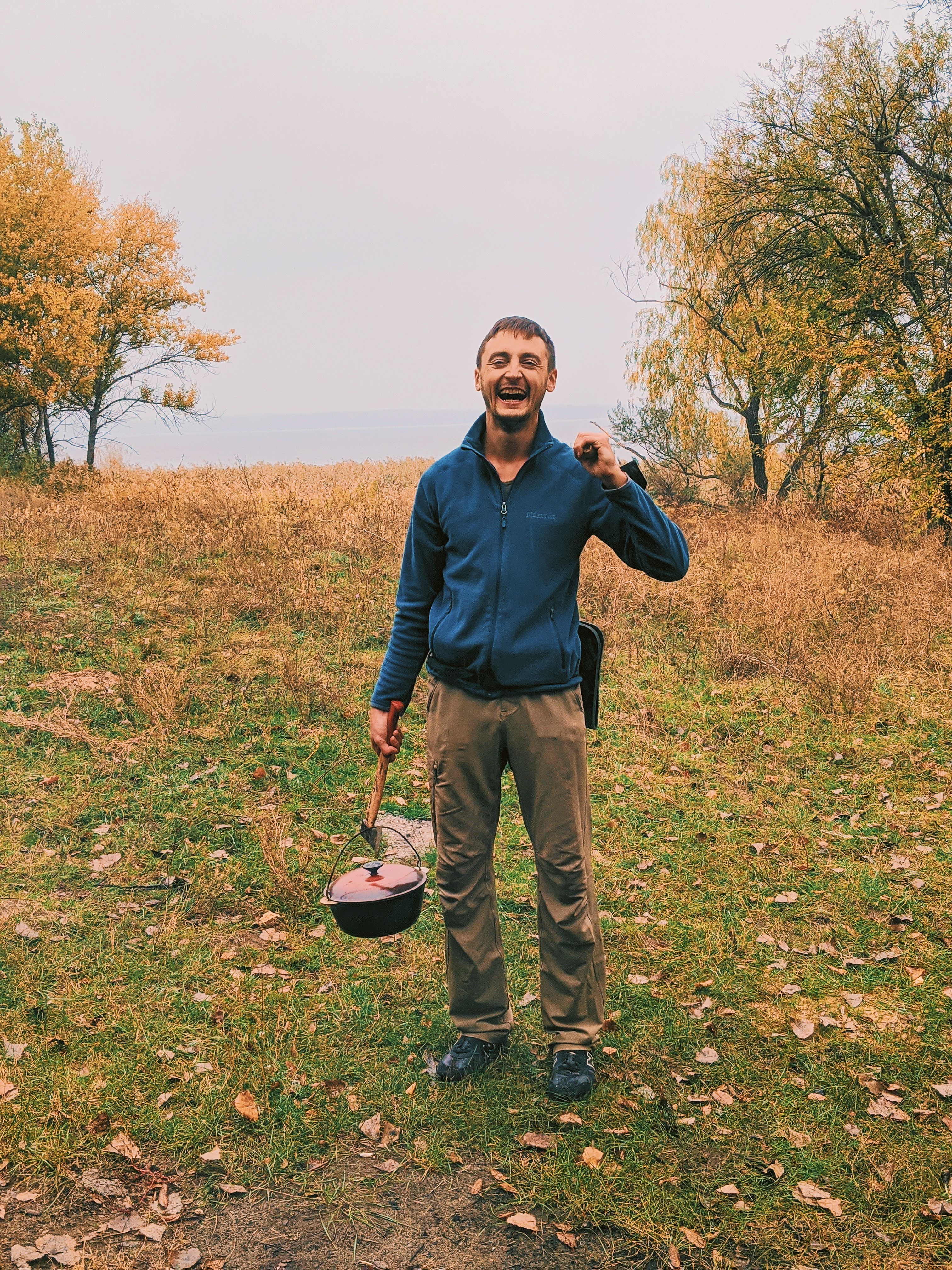 Full body of smiling male camper standing with pot and tool on grassy ground with trees during camping in woods