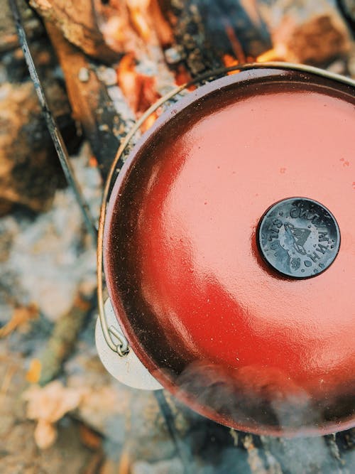 Top view of red metal pot with lid placed on burning firewood while cooking in nature on blurred background during camping