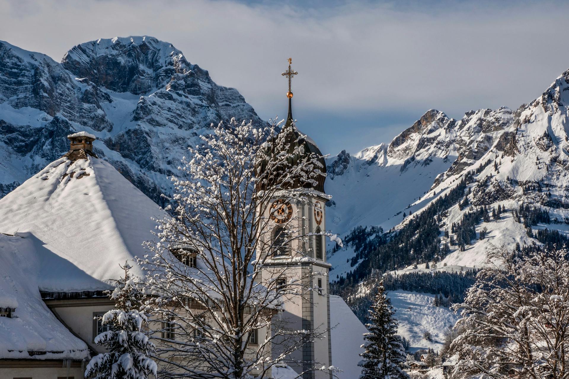 Exterior of stone church with black dome near snowbound houses and leafless trees near mountain ridge under blue gray sky