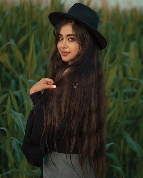 A Woman with Long Hair Wearing Black Fedora Hat Standing Near Tall Grass while Posing at the Camera