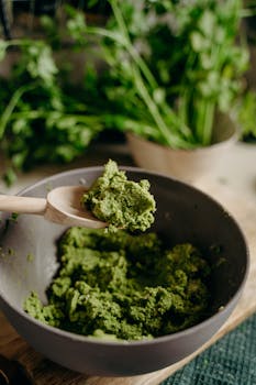 A close-up of fresh green pesto in a ceramic bowl with a wooden spoon, set in a rustic kitchen.