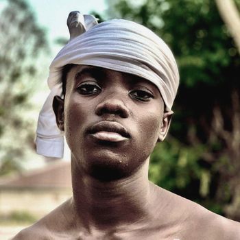 A serious young boy with a headscarf stands outdoors, conveying a strong and thoughtful expression.