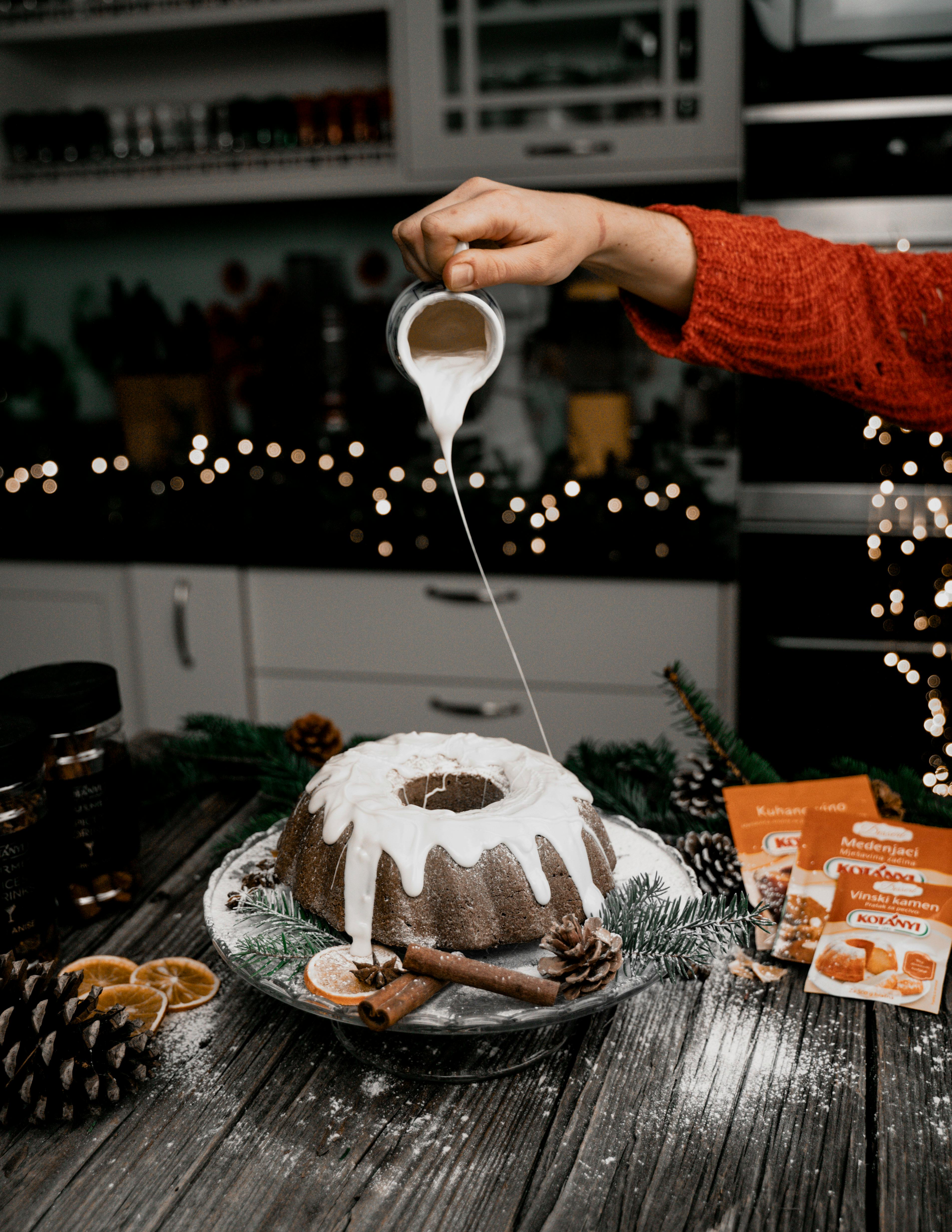 Hand pouring sugar icing over a bundt cake in a festive decorated kitchen setting.